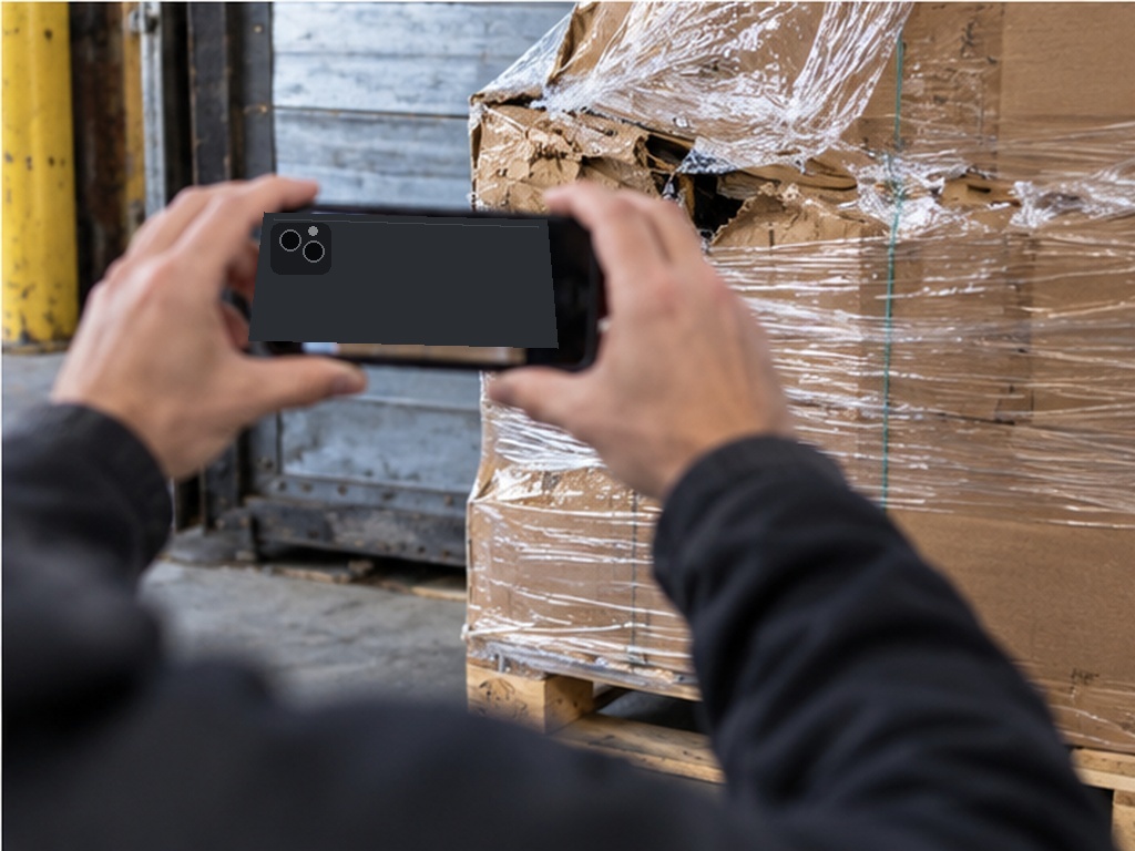 Two hands holding a phone up in landscape to photograph a stretch-wrapped pallet with a torn and crushed carton at its corner, at a loading dock.