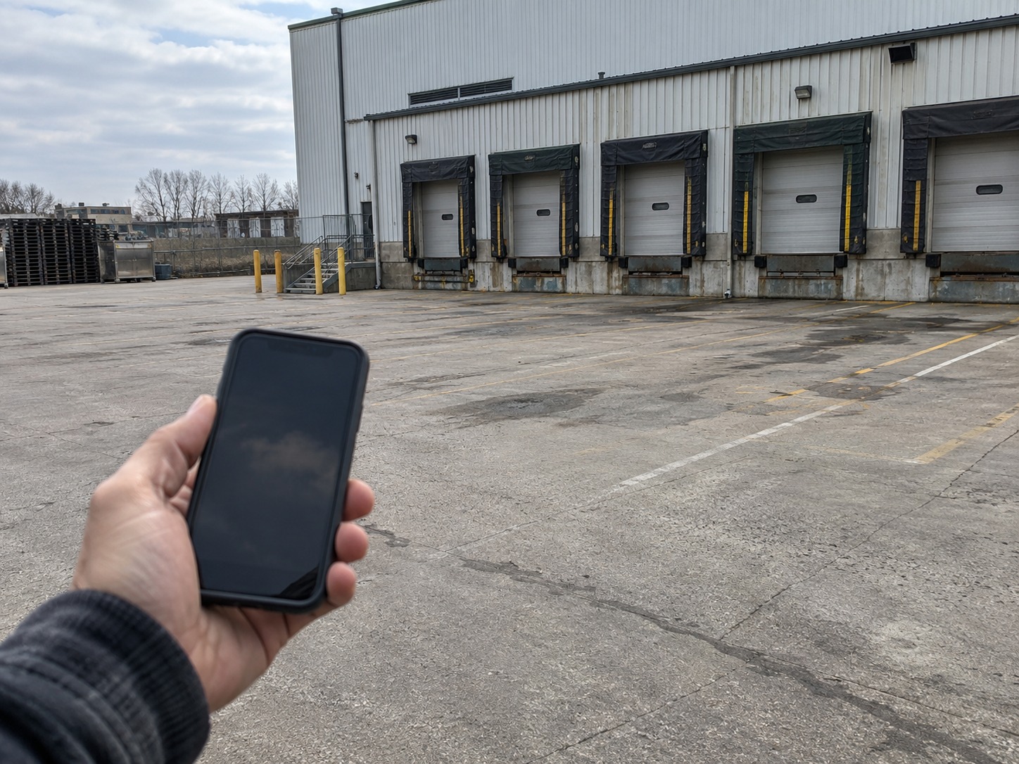 A hand holding a phone with a blank screen in the foreground, looking out across an empty dock apron at a row of closed dock doors with no truck present.