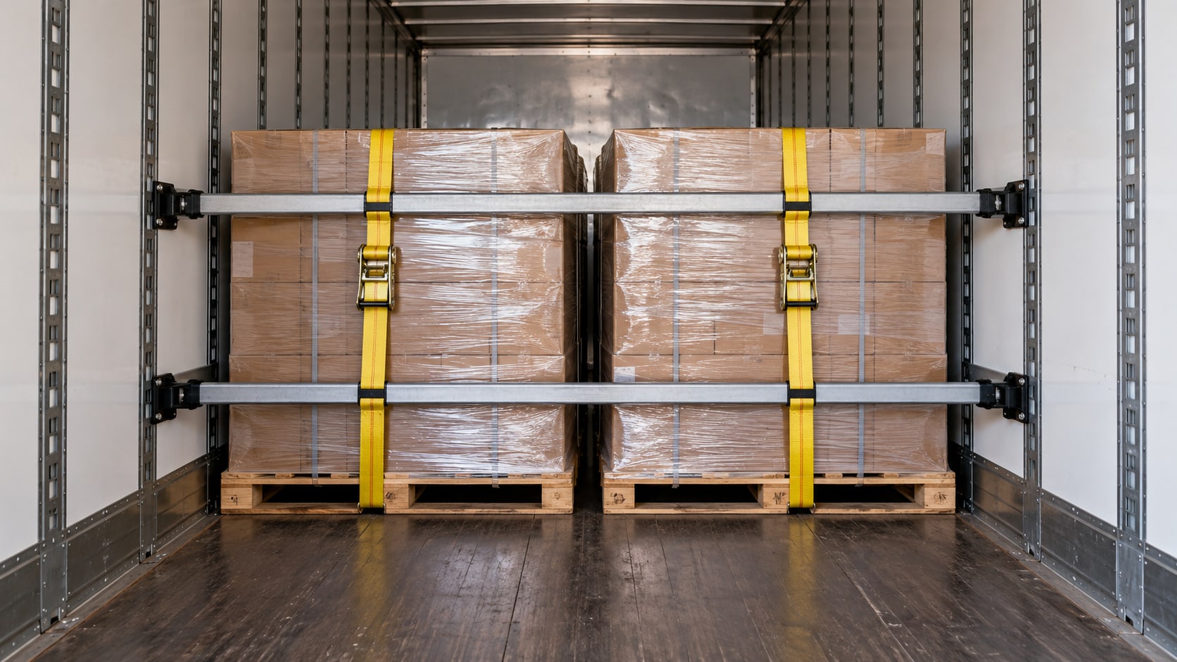 Two pallets inside a trailer held by load bars and yellow ratchet straps anchored to the logistics track.