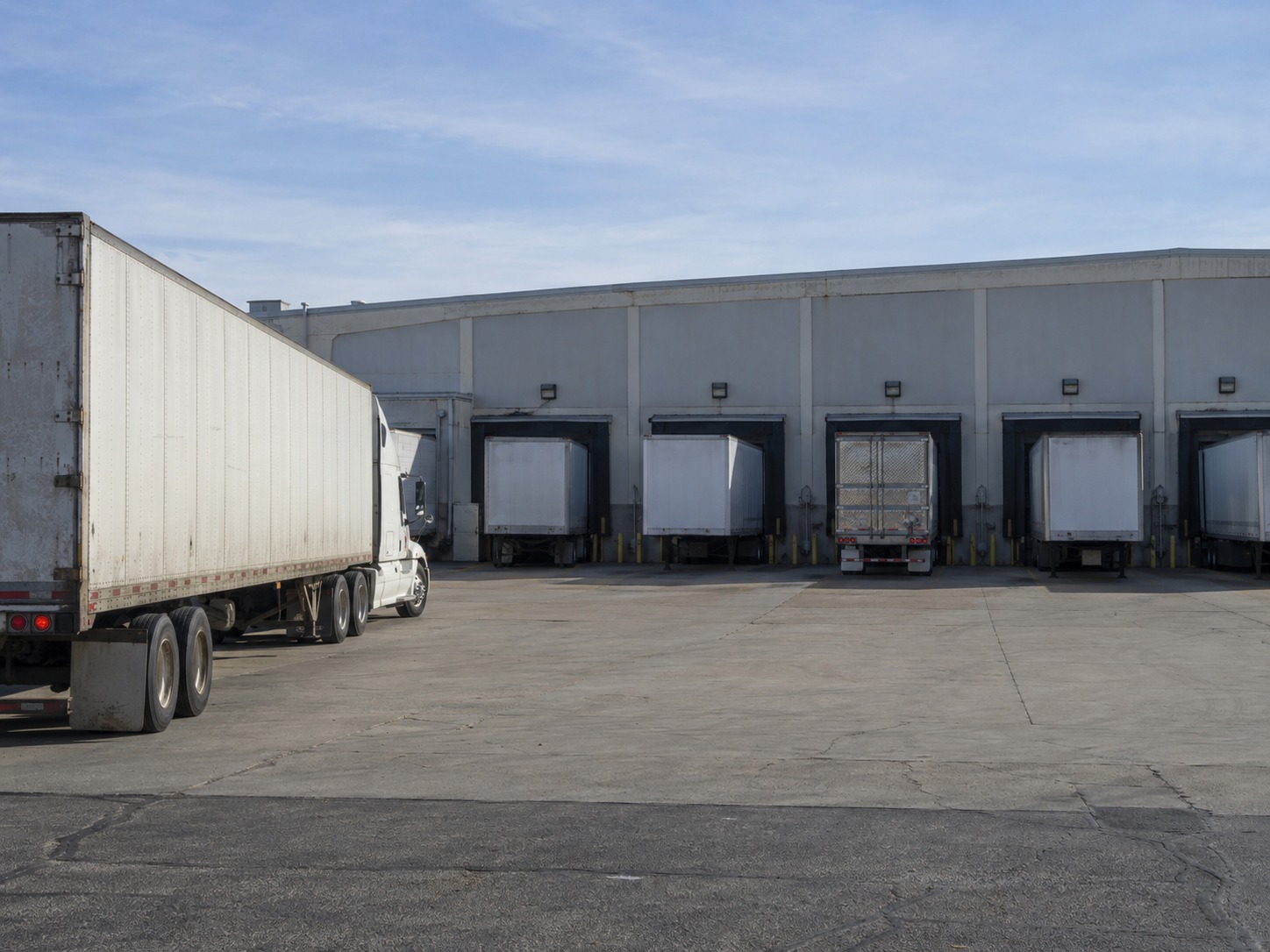 A tractor-trailer parked and waiting on a yard apron while every dock door along the building ahead is already occupied by other trailers.