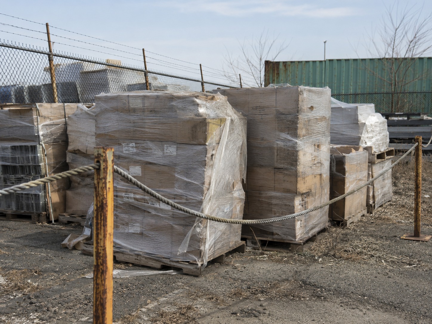 Several stretch-wrapped pallets behind a rope barrier in an outdoor yard, the wrap and cartons weathered from sitting outside.